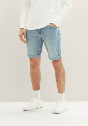 Homme portant un short en jean bleu clair retroussé, une chemise blanche à manches longues, des chaussettes blanches et des baskets blanches, debout devant un fond uni.