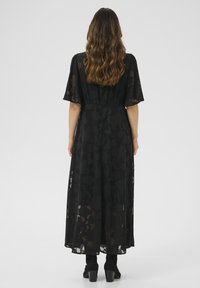 Vestito maxi nero con sovrapposizione trasparente, maniche corte e motivo floreale. Il vestito ha una vita arricciata e scorre lungo fino alla caviglia, indossato con stivaletti neri.