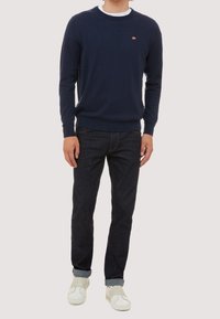 Pull en coton bleu marine à col rond, porté sur un T-shirt blanc. Associé à un jean foncé et des baskets blanches, avec les poignets retroussés.