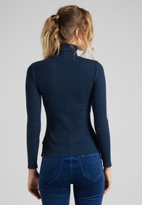 Haut à manches longues bleu marine avec un col montant, présentant une texture côtelée verticale et de subtiles rayures marron. Associé à un pantalon en velours côtelé bleu.