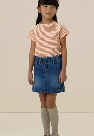 Jeune fille aux cheveux noirs raides portant un t-shirt à rayures orange et blanches, une jupe en jean et des chaussettes beige montant jusqu'aux genoux, debout devant un fond uni.