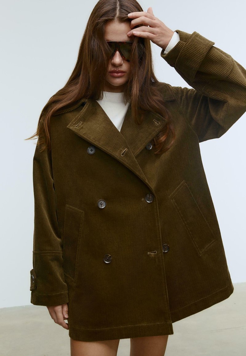 Brauner corduroy Doppelreiher-Mantel mit weitem Kragen, großen Knöpfen, seitlichen Taschen und oversized Passform. Das Model trägt Sonnenbrillen und ein weißes Oberteil.