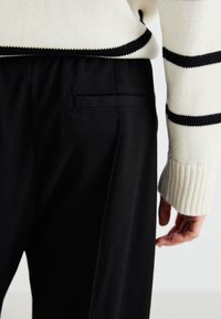 Sweter dzianinowy w kremowym kolorze z czarnymi paskami, zestawiony z czarnymi spodniami z jedną tylną kieszenią i teksturowanym materiałem.