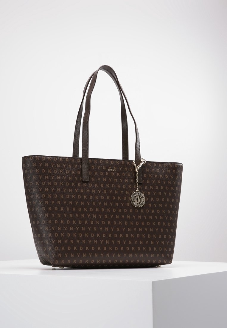 Sac fourre-tout DKNY marron foncé avec motif de logo répété, deux bretelles, et un pendentif logo argenté suspendu, posé sur une surface blanche.