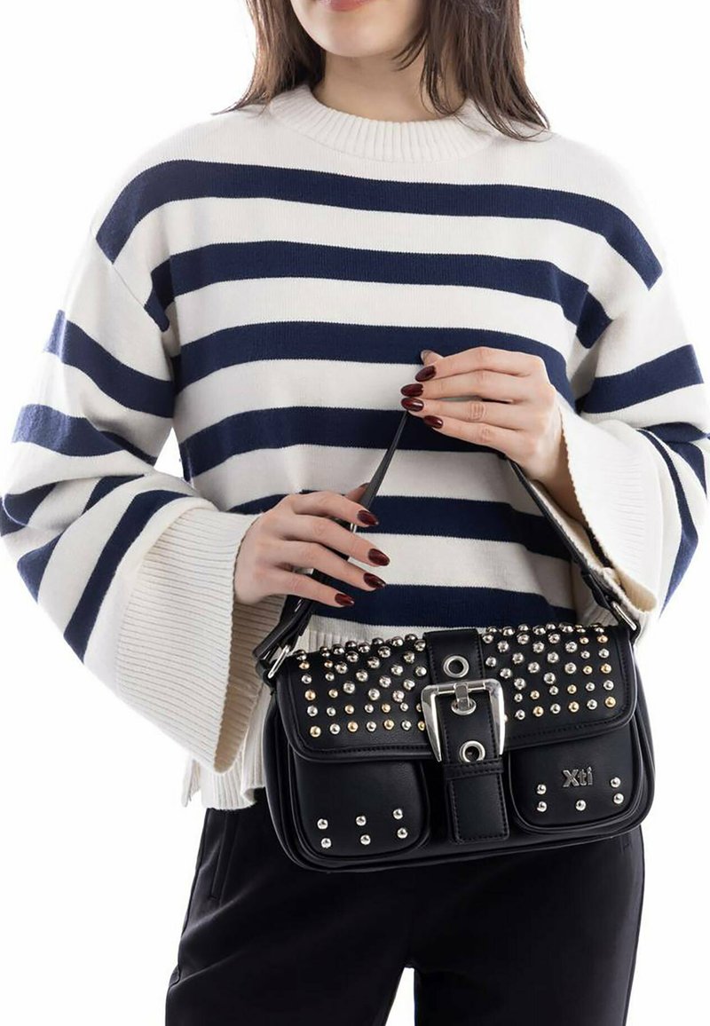 Mujer vistiendo un suéter a rayas blancas y marinas sosteniendo un bolso negro con tachuelas, con una hebilla y el logo "Xti".