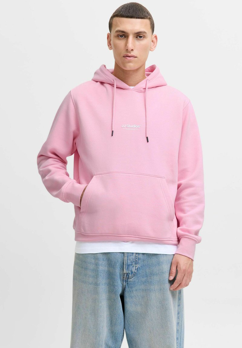Różowa bawełniana bluza z kapturem, z troczkiem, przednią kieszenią i białym logo "JJ Studios". W połączeniu z jasnoniebieskimi dżinsami o luźnym kroju.