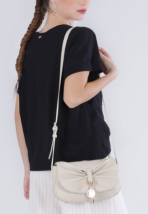 Mujer con cabello largo trenzado, vestida con un top negro y pantalones blancos con rayas, llevando un bolso cruzado beige con un detalle de anillo dorado.