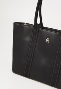 Bolso tote negro hecho de material liso, con dos asas superiores, detalle de logo dorado y correas laterales tejidas con el nombre de la marca.