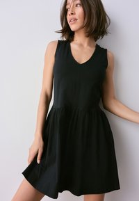 Sort ærmeløs kjole med V-hals, tætsiddende overdels og udvidet nederdel; lavet af blødt stof med en glat tekstur og et minimalistisk design.