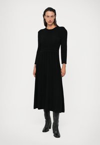 Vestido preto de manga longa em malha com corpete justo e saia rodada, apresentando pregas verticais e uma textura suave, combinado com botas pretas até ao joelho.