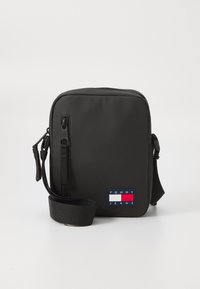 Black rectangular messenger bag made of smooth material. Features a shoulder strap and a front patch with the "TOMMY JEANS" logo in red, white, and blue.