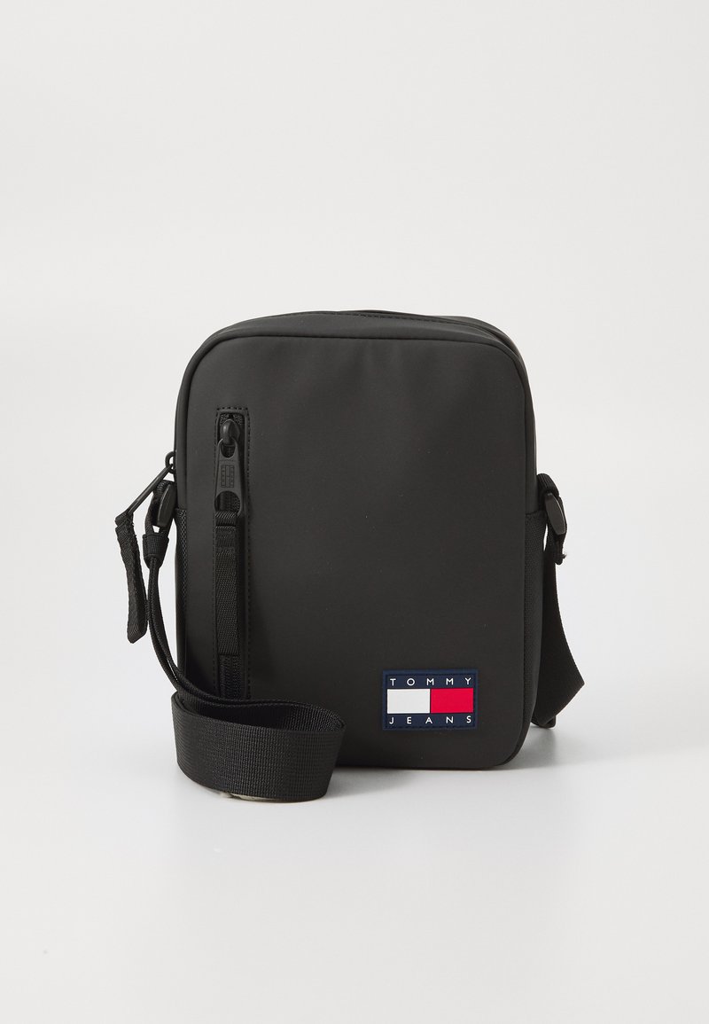 Borsa a tracolla rettangolare nera realizzata in materiale liscio. Presenta una tracolla e una patch frontale con il logo "TOMMY JEANS" in rosso, bianco e blu.