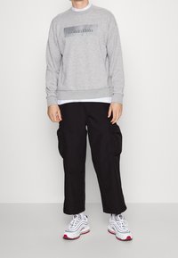 Šedá mikina s lesklým logem Calvin Klein, doplněná volnými černými cargo kalhotami s bočními kapsami a teniskami s bílými prvky a červenými akcenty.