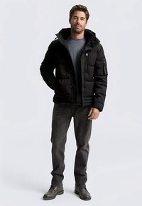 Zwart pufferjack met een ritssluiting aan de voorkant en drukknopen, voorzien van een afneembare capuchon en een zak met rits op de mouw, gecombineerd met donkere jeans en laarzen.