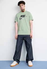 Zelené bavlněné tričko s černým logem Nike, spárované s tmavě modrými volnými džíny a bílými teniskami s gumovou podešví.