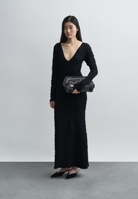 Langes schwarzes Kleid mit strukturierter Stoff, tiefem V-Ausschnitt und langen Ärmeln, kombiniert mit einer Clutch mit aufwendigen Metallelementen.