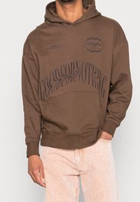 Felpa marrone in morbido cotone con un design testuale grafico, grande tasca a marsupio e polsini a costine. Abbinata a pantaloni a righe chiare.