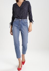 Blusa navy con stampa floreale bianca, maniche lunghe con fiocchi, abbinata a jeans slim-fit azzurri chiari e scarpe con tacco rosso.