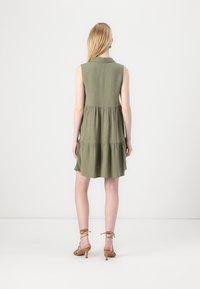Robe sans manches verte avec un col boutonné, des volants superposés et une coupe décontractée. Le tissu a une texture subtile. Sandales brunes à talons.