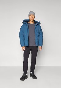 Blå vadderad jacka med huva, bärs över en grå T-shirt, kombinerad med svarta byxor och svart skor. Modellen står mot en neutral bakgrund.