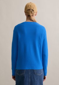 Jasnoniebieski sweter z długimi rękawami i okrągłym dekoltem, wykonany z gładkiej tkaniny. W połączeniu z dżinsową spódnicą, pokazany z prostego tyłu.