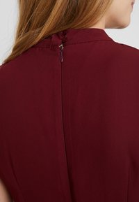 Burgundské šaty s bezešvým designem, s viditelným zipovým uzávěrem na zadní straně a hladkým, jemným textilním povrchem.