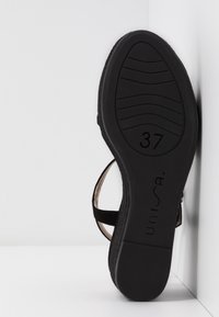 Semelle de sandale noire taille 37 avec le nom de marque Unisa visible, positionnée debout contre un mur d'angle blanc.