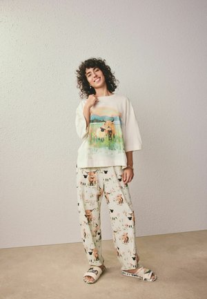 Kvinde smilende i cremefarvet pyjamasæt med farverige ko- og blomsterprint, stående foran en tekstureret beige væg og gulv.