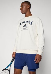 Krēmveida džemperis ar "ADIDAS" un "TENNIS CLUB" tekstu jūras zilā krāsā, ribots piedurkņu un apakšmalas dizains. Modelis valkā jūras zilas šortus un tur rokas tenisa raketi.