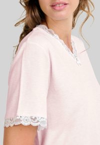 Mujer vistiendo una blusa de manga corta color rosa claro con borde de encaje blanco en el escote en V y en los bordes de las mangas, y un pequeño detalle de lazo en el collar.