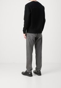 Osoba w czarnej bluzie, szarych dżinsach i czarnych skórzanych butach, stojąca tyłem na jednolitym białym tle.