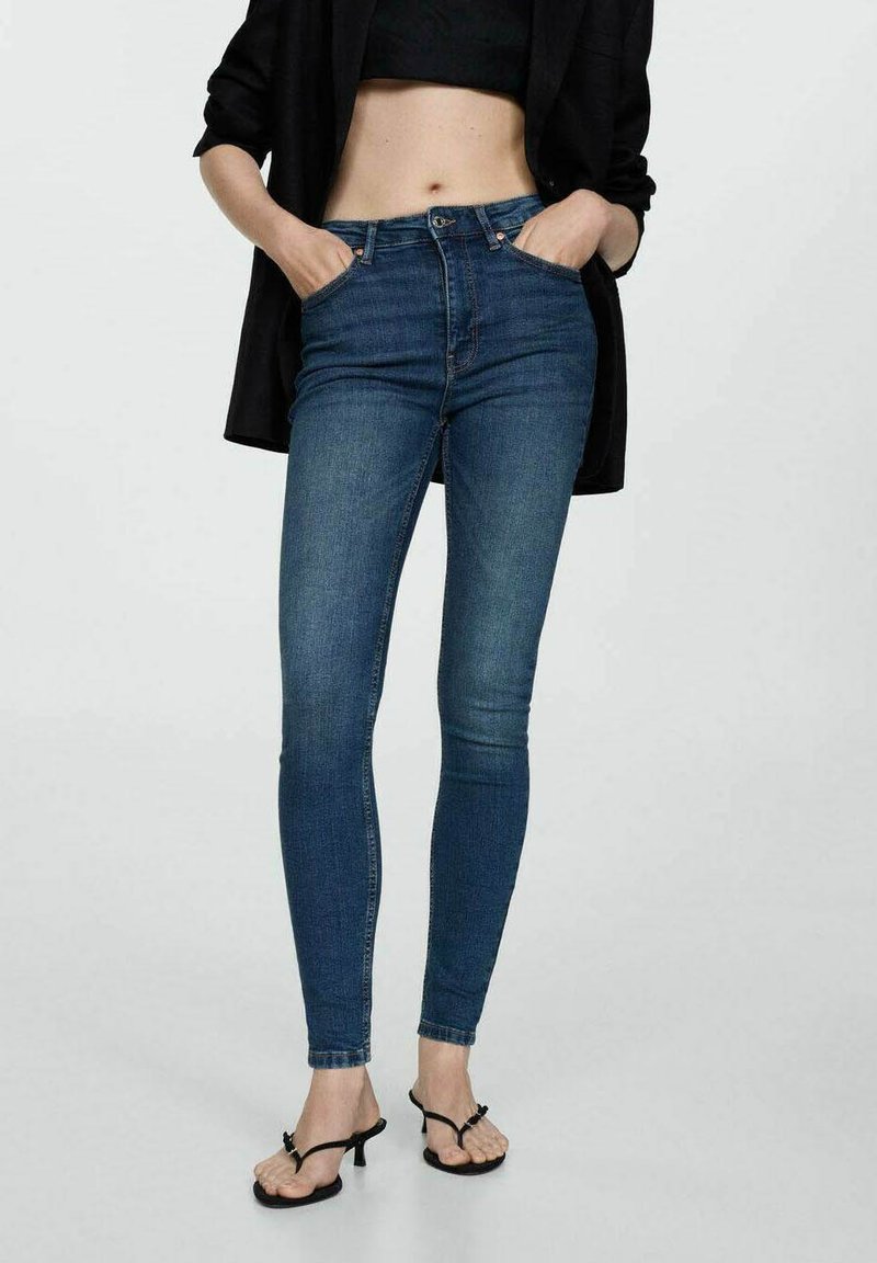 Oprijete kavbojke v temno modrem denimu, z visokim pasom in sprednjimi žepi, v kombinaciji s črnimi sandaali. Tekstura je videti gladka in prilegnjena.