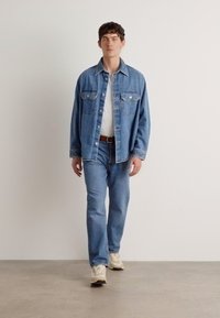 Denimjakke med to brystlommer, lyseblå jeans og kremfargede sneakers. Modellen står mot en ensfarget bakgrunn, med avslappede armer.