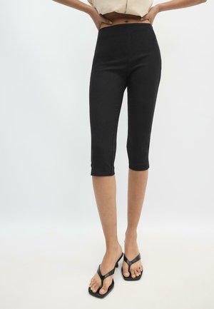 Žena nosi čierne priliehavé krátke legíny a čierne sandále na podpätku, ruky opreté na bokoch na jednofarebnom svetlom pozadí.