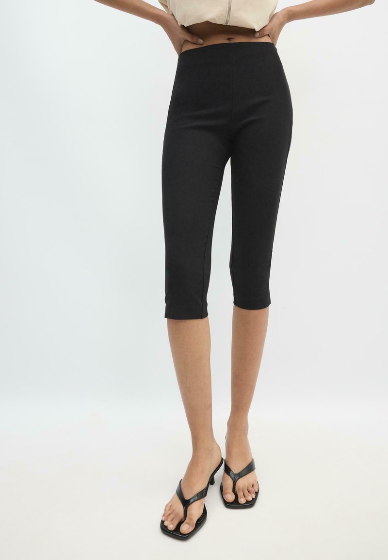 Femme portant des leggings noirs ajustés courts et des sandales noires à talons, mains posées sur les hanches sur un fond clair uni.
