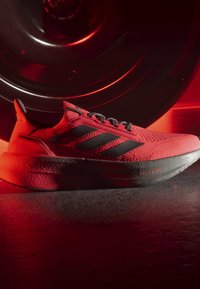 Chaussure de course rouge avec des bandes noires et des lacets gris, positionnée devant une grande roue noire éclairée en rouge en arrière-plan.