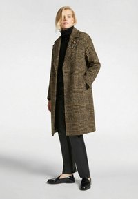 Manteau en laine à carreaux marron et noir avec un design croisé et un col à revers, porté avec un col roulé noir et un pantalon noir.