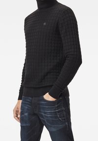 Svart stickad tröja med ett fyrkantigt mönster, hög krage och ribbade ärmslut och fåll. Matchad med mörkblå denimjeans.