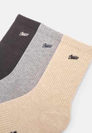 Tre par ribbede sokker i mørkebrun, grå og beige med et logo på kanten. De har et sømløst tådesign og en blød tekstur.