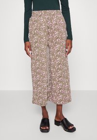 Calças largas com padrão, num fundo castanho com um design floral rosa; cintura elástica; usadas com sapatos pretos de deslizar.