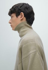 Beiger Rollkragenpullover aus weichem Strick mit geripptem Kragen, seitlichen Nahtdetails und übergroßer Passform, getragen von einem nach links blickenden Model.