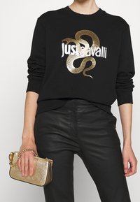 Svart sweatshirt med guldorm-grafik och vit text, i kombination med en strukturerad guldclutch med kedjesnodd. Svarta läderbyxor.