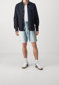 Camisa de denim negra, abotonada con un pequeño logo; combinada con pantalones cortos claros, remangados, y zapatillas blancas con acentos negros.