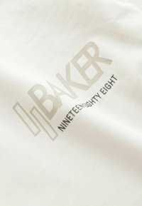 Tissu blanc avec le texte stylisé "H BAKER" en beige clair et un texte plus petit en noir indiquant "NINETEEN EIGHTY EIGHT" imprimé dessus.