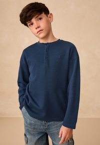 Garām piedurknēm tumši zils ribots henlē kreklā ar trim pogām. Krekls ir ar apaļu kakla izgriezumu un nelielu logotipu uz kreisā krūtīs.