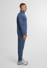 Gaiši zils sporta kostīms ar augstu apkakli, ar divām tumšām svītrām pa piedurknēm un atbilstošām biksēm. Strukturēta auduma un pieguļošs dizains.