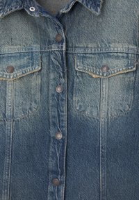 Chaqueta de mezclilla azul claro con una textura desgastada, bolsillos en el pecho dobles y cierre de botones. Cuenta con botones de metal y un collar puntiagudo.