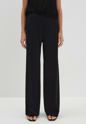 Zwarte wijde broek van een zachte, vloeiende stof. Heeft een hoge taille en een minimalistisch ontwerp, gecombineerd met een doorschijnende, getextureerde top.