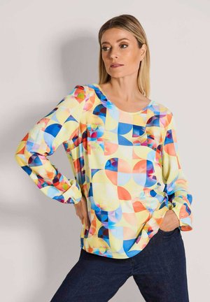 Frau trägt eine langärmelige Bluse mit mehrfarbigen geometrischen Formen und dunkelblaue Jeans, steht mit einer Hand in der Tasche.