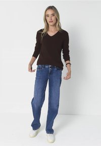 Hnědý svetr s výstřihem do V, modré rovné džíny a bílé tenisky. Materiál je hladký s přiléhavým střihem a jednoduchým stylem.